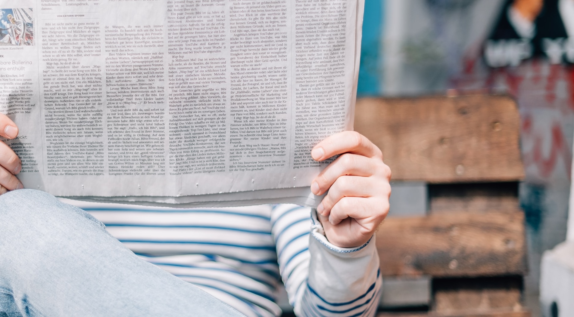 Closely cropped photo of a man holding a newspaper in front of his face, sitting on a park bench