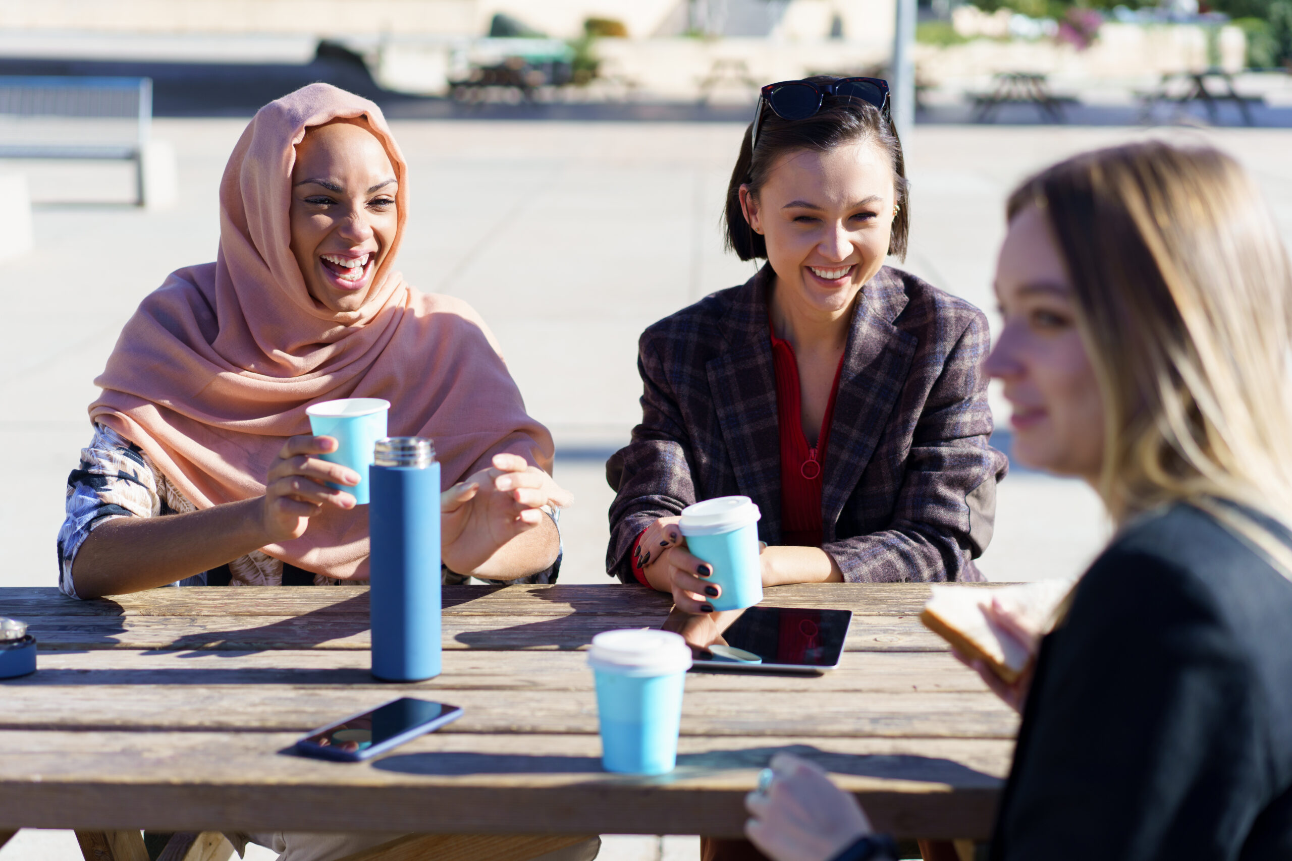 Happy young diverse female friends, in casual clothes, smiling while resting in outdoor cafe and drinking coffee from take-away cups