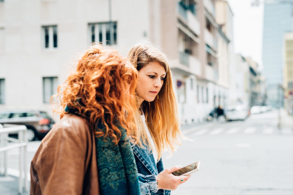 Two young women-identifying friends strolling outdoors in the city with smart phone