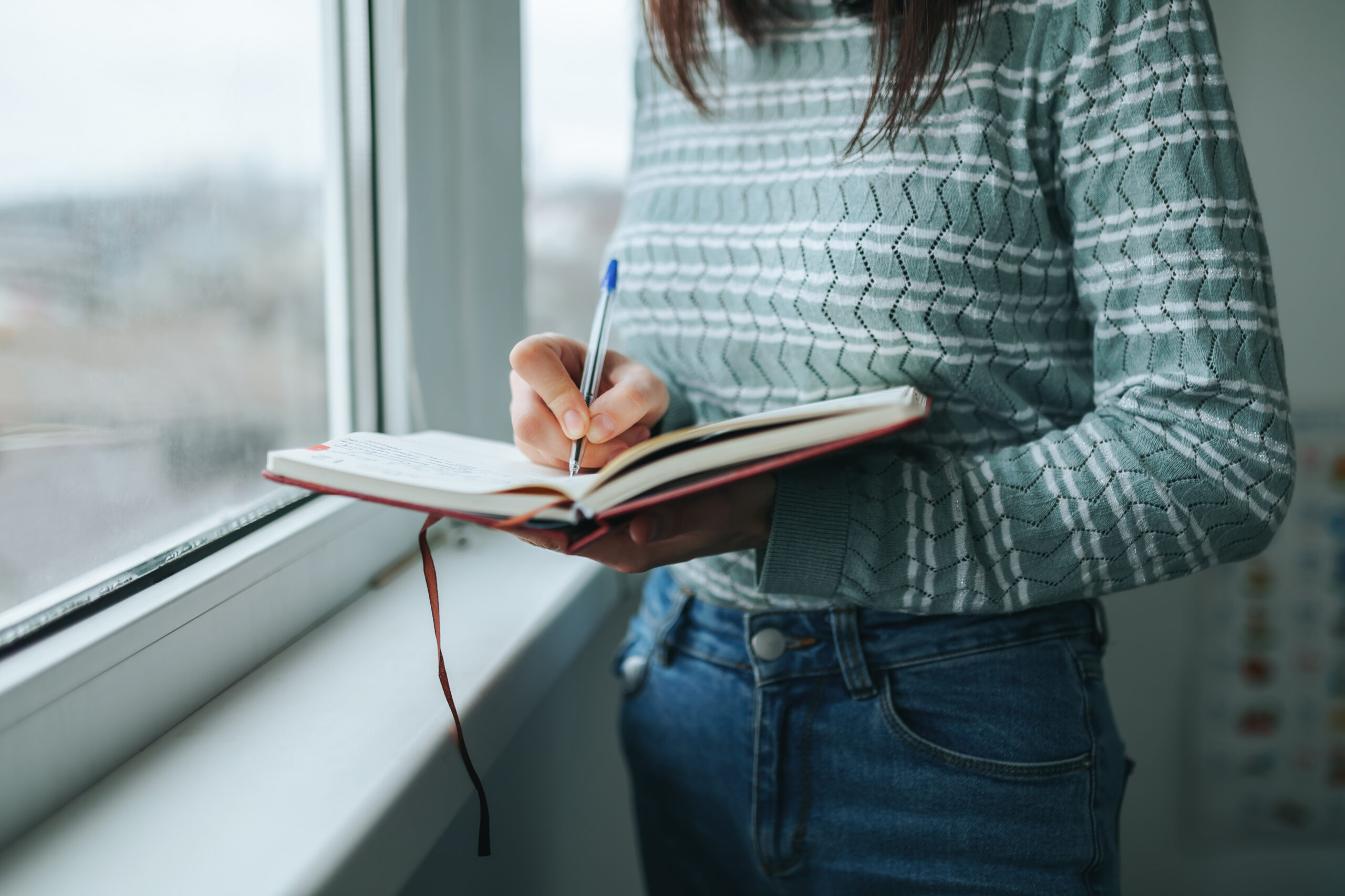 Woman-presenting figure stands at a window and writes with a pen in a notebook.