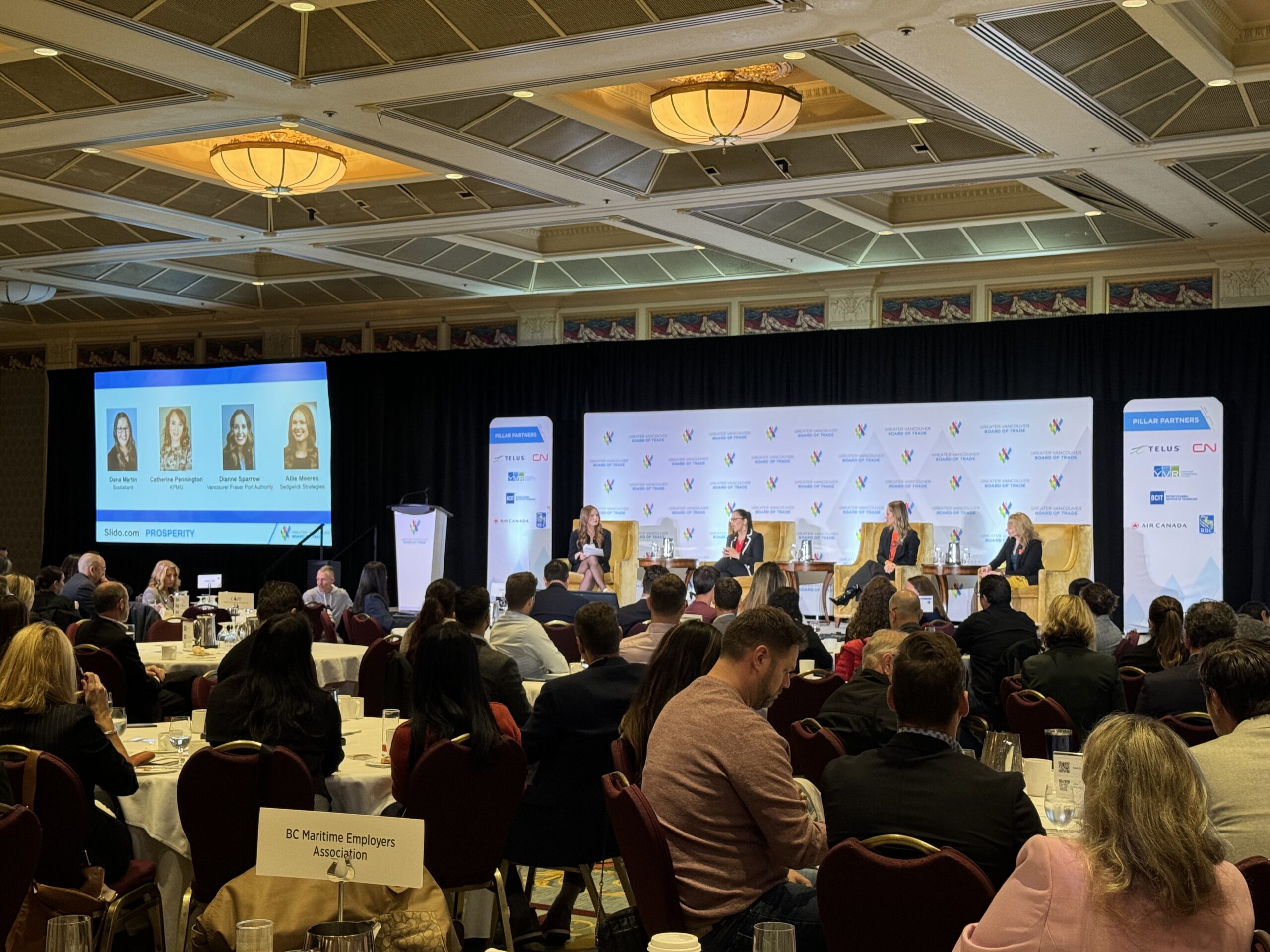 four women sit on chairs on a stage in a hotel ballroom for a panel discussion