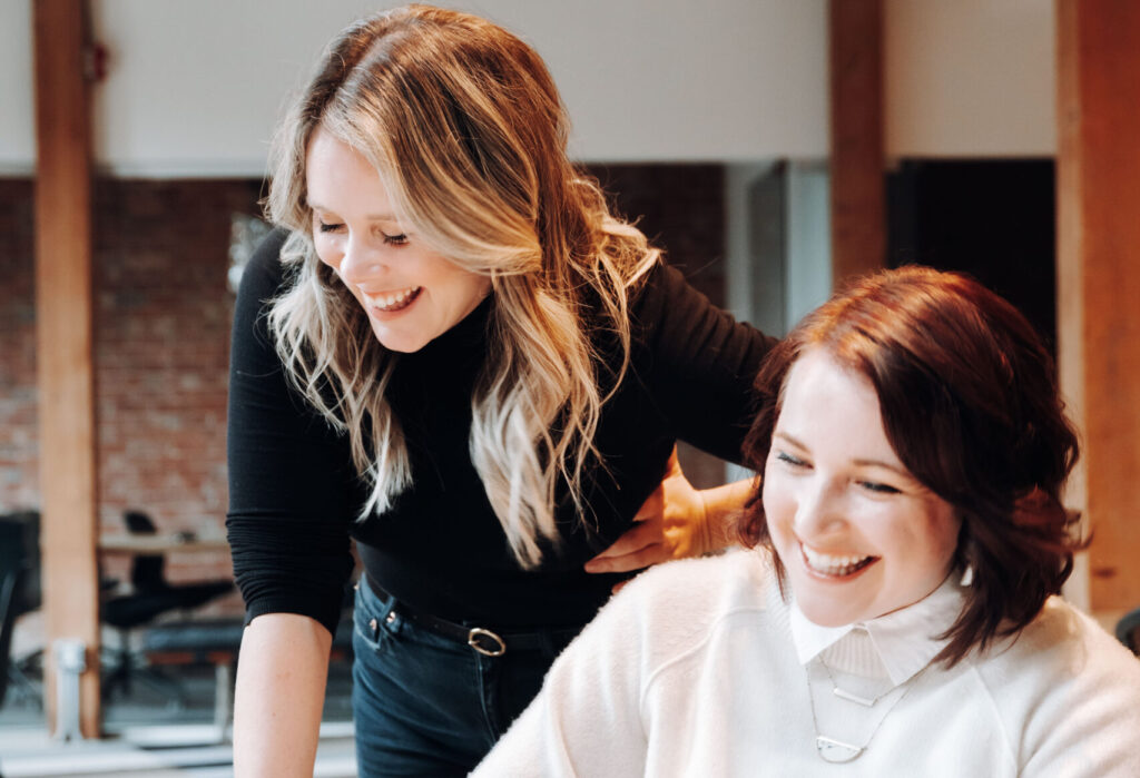 two women laugh next to each other, one with blonde hair and a black turtleneck and the other with auburn hair and a white sweater