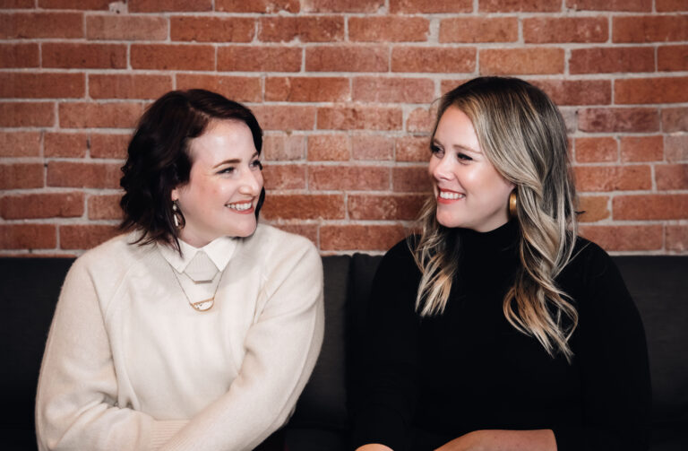 one auburn haired women in a white sweater looks at another woman with blonde hair and a black turtleneck