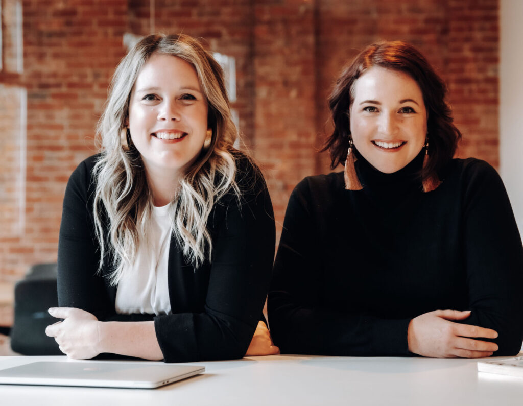 two women look at the camera and lean on a table. one has blonde hair and a black blazer, the other has auburn hair and a black turtleneck