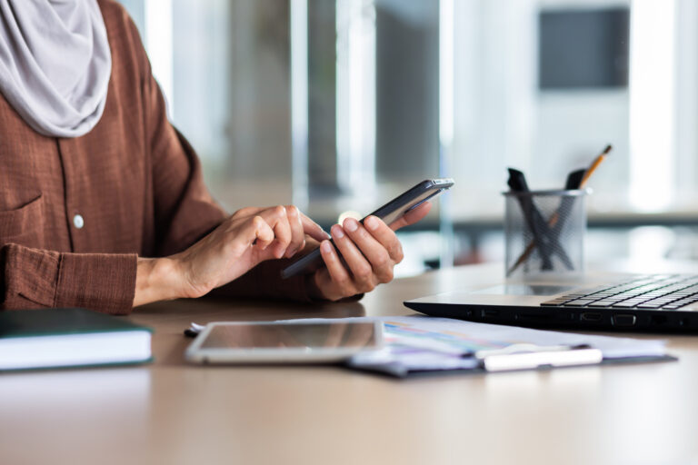 Close up hands of a woman of colour use the phone, with a computer and notebooks nearby on her desk
